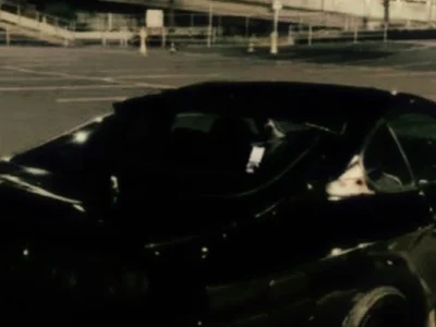 A darkly lit image of a parked black car in an empty parking lot at night.
