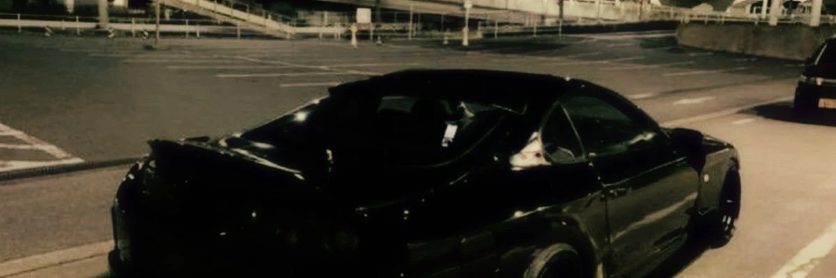 A darkly lit image of a parked black car in an empty parking lot at night.